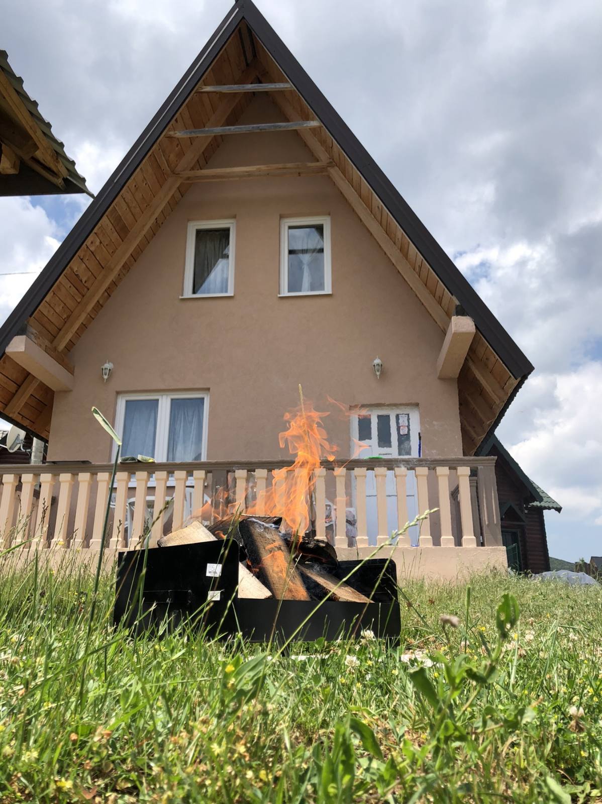 A charming house with a pitched roof is set against a backdrop of clouds. In the foreground, a fire pit with flames flickers, surrounded by green grass. The house features two large windows and a welcoming porch with a wooden railing.