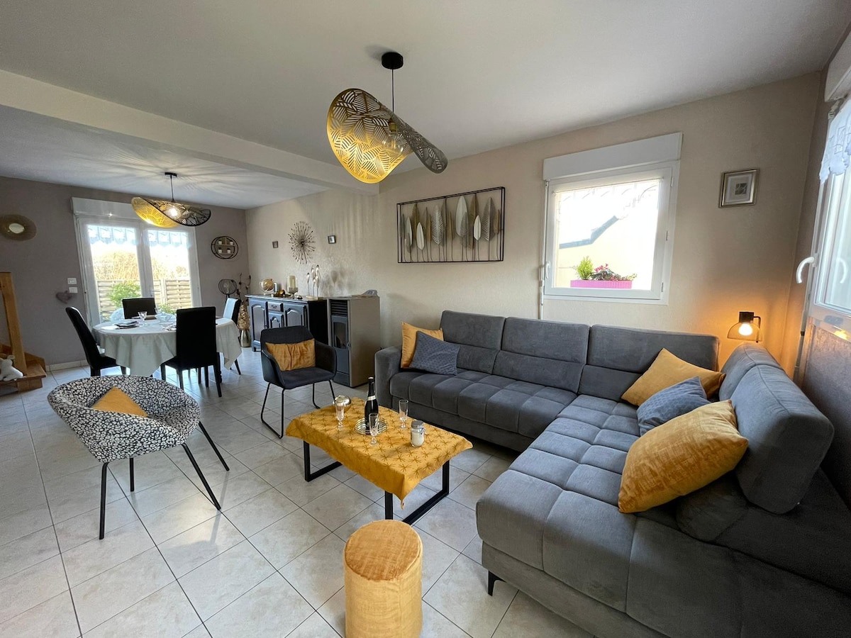 A spacious living area features a comfortable gray sectional sofa with vibrant yellow cushions. A coffee table is centrally located, complemented by decorative elements. In the background, a dining area is visible with a white table and chairs, along with large windows allowing natural light to fill the room.