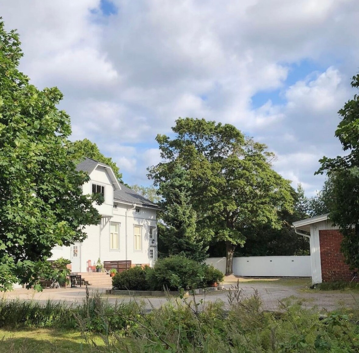 A charming old wooden house is surrounded by greenery, featuring a large tree and well-maintained shrubs. The building showcases a light exterior with a staircase leading to a patio area, creating a serene outdoor space. A red brick garage is visible to the side.