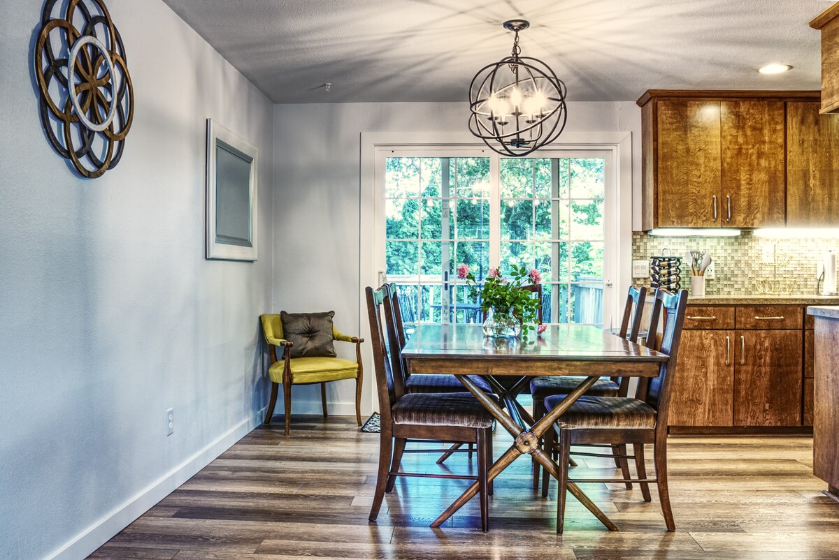 A dining area features a wooden table surrounded by six chairs and accented by a decorative light fixture. A bouquet of flowers sits in the center of the table. Large windows allow natural light to fill the space, offering a view of the outdoors.