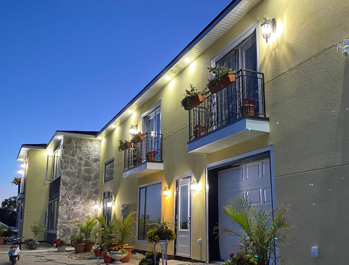The exterior of the villa is illuminated in the evening light, highlighting a modern structure with stone accents. Flower baskets adorn the balconies, and various potted plants line the entrance, contributing to a welcoming environment.