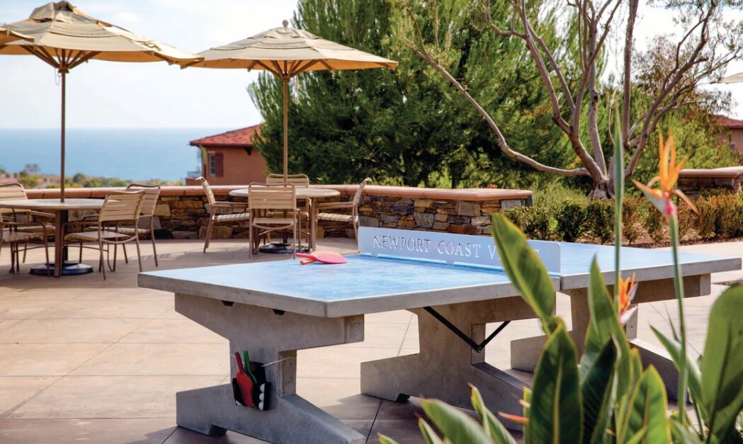 An outdoor area features a ping pong table, surrounded by neatly arranged tables and chairs under shade umbrellas. Lush greenery and low plants are visible in the foreground, while the serene ocean can be seen in the background, contributing to a relaxing atmosphere.