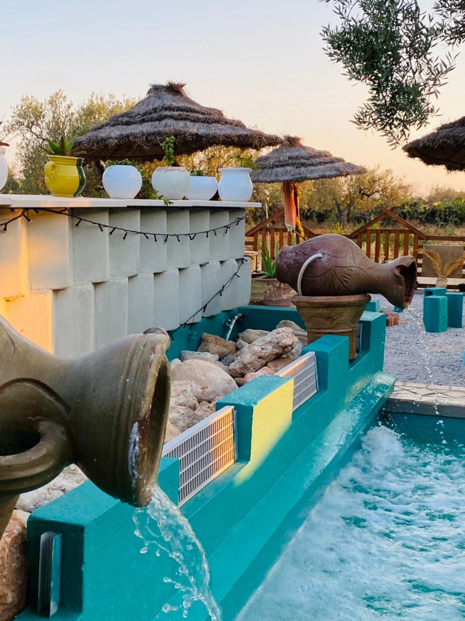 Decorative clay pots channel water into a turquoise pool, surrounded by natural stone and greenery. Thatched umbrellas offer shade in the background, while potted plants add a touch of color along the wall, creating a peaceful outdoor environment.