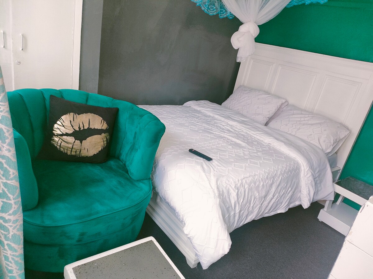 A cozy sleeping area features a spacious bed covered with crisp white linens. A plush teal armchair is positioned nearby, adorned with a decorative pillow. Light green walls complement the overall design, while natural light enhances the inviting atmosphere.