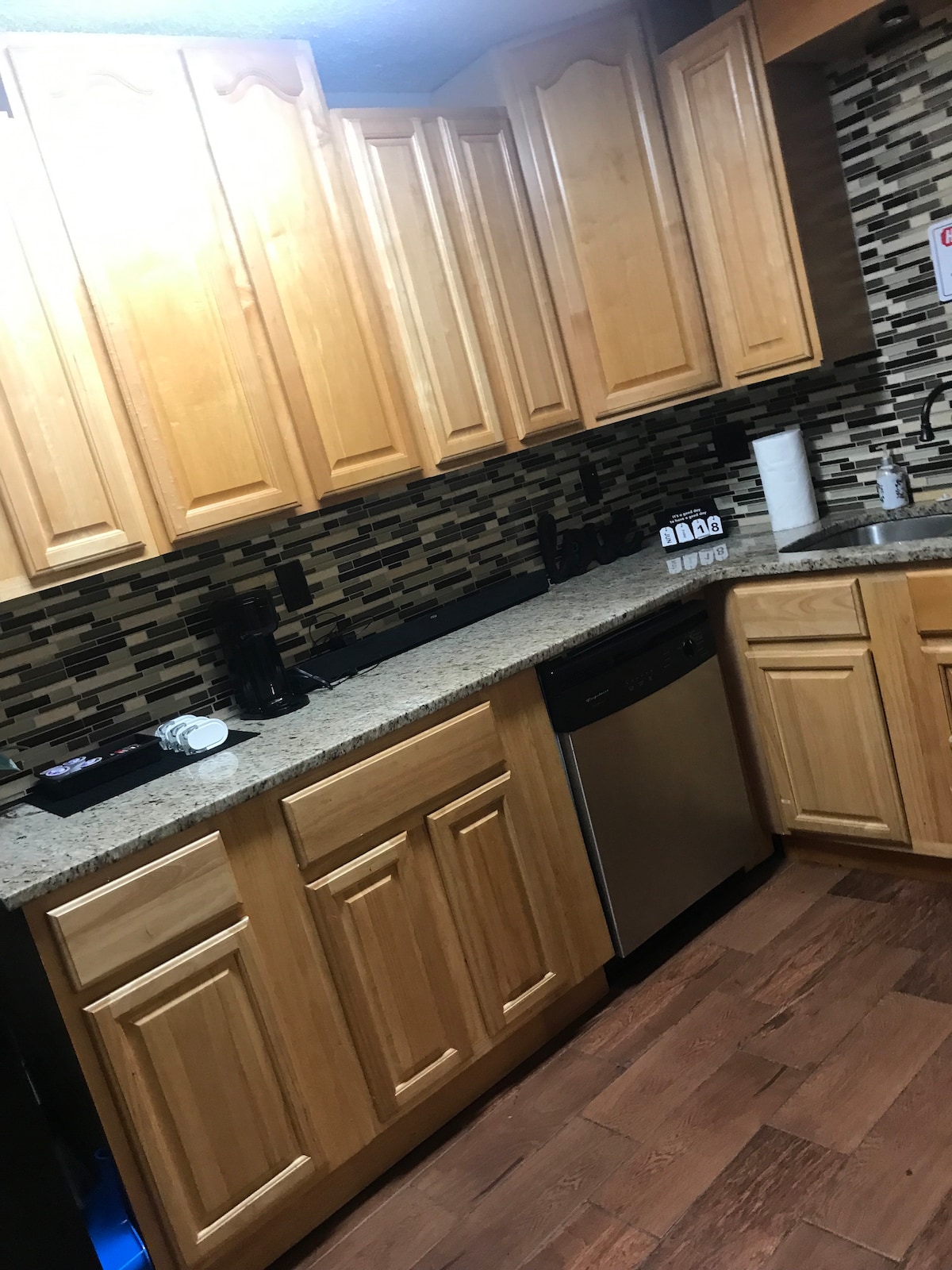 The kitchen features cabinetry in warm wood tones, paired with a speckled granite countertop. Modern appliances, including a dishwasher and coffee maker, are visible. A backsplash made of dark tiles adds a stylish contrast to the space, complemented by wood-like flooring.