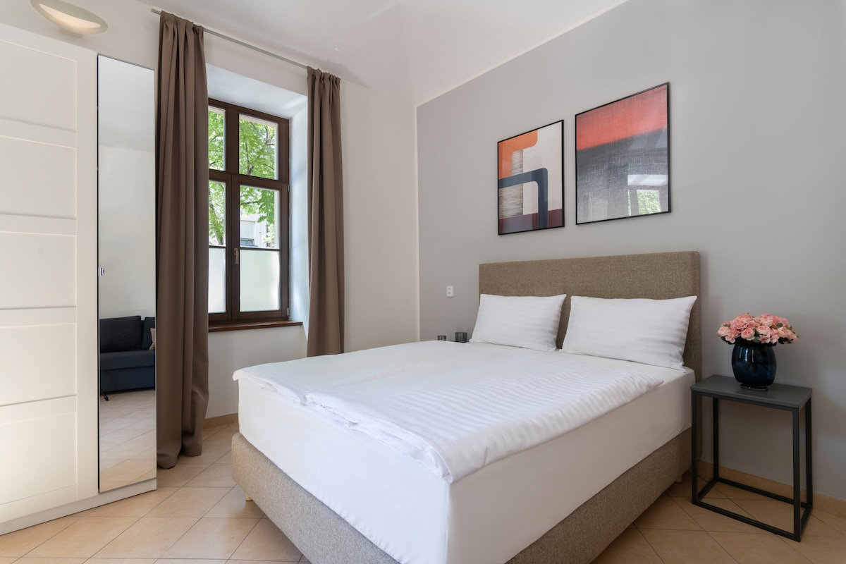 A spacious bedroom features a comfortable bed dressed with crisp linens. Nearby, a sleek nightstand holds a vase of flowers. Natural light filters through a large window adorned with elegant curtains, creating a bright and inviting space.