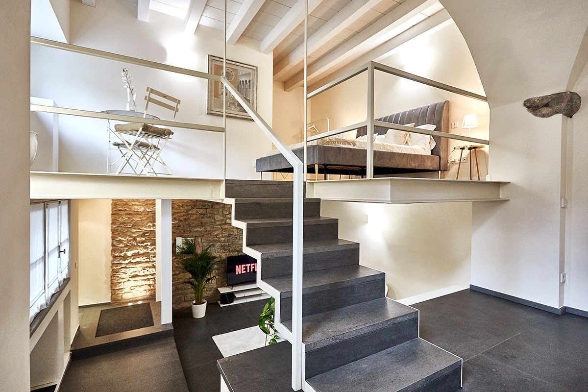 A spacious loft is depicted, featuring a charming split-level design. A staircase leads to an upper area containing a cozy bed framed by white beams. Below, a stylish seating area with a small table and chairs offers a welcoming space, complemented by a stone wall.