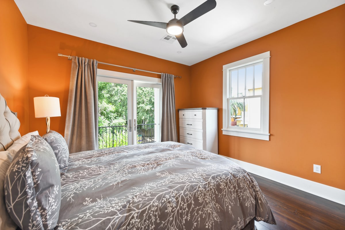 A warm-toned bedroom is highlighted by orange walls and features a large bed with floral-patterned bedding. Natural light enters through double windows covered by light curtains. A modern ceiling fan is installed above, and a dresser stands against the wall near the windows.