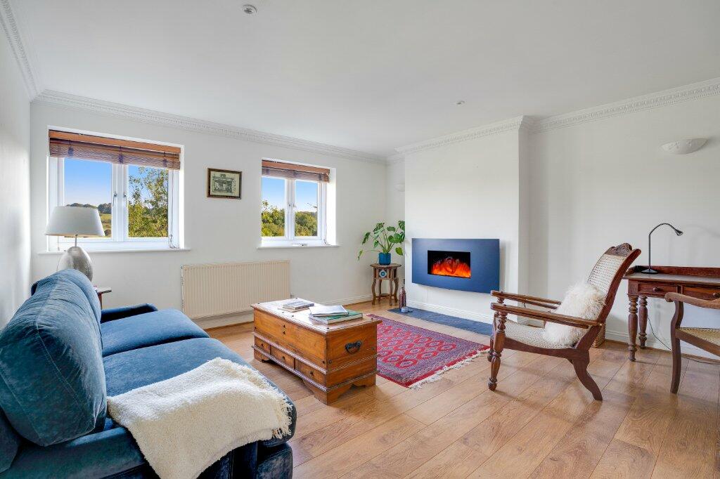 A bright living room features large windows revealing valley views. A comfortable blue sofa is complemented by a wooden coffee table and a rustic accent chair. An electric fireplace provides warmth, while a small writing desk adds functionality. Light wooden flooring enhances the spacious feel.