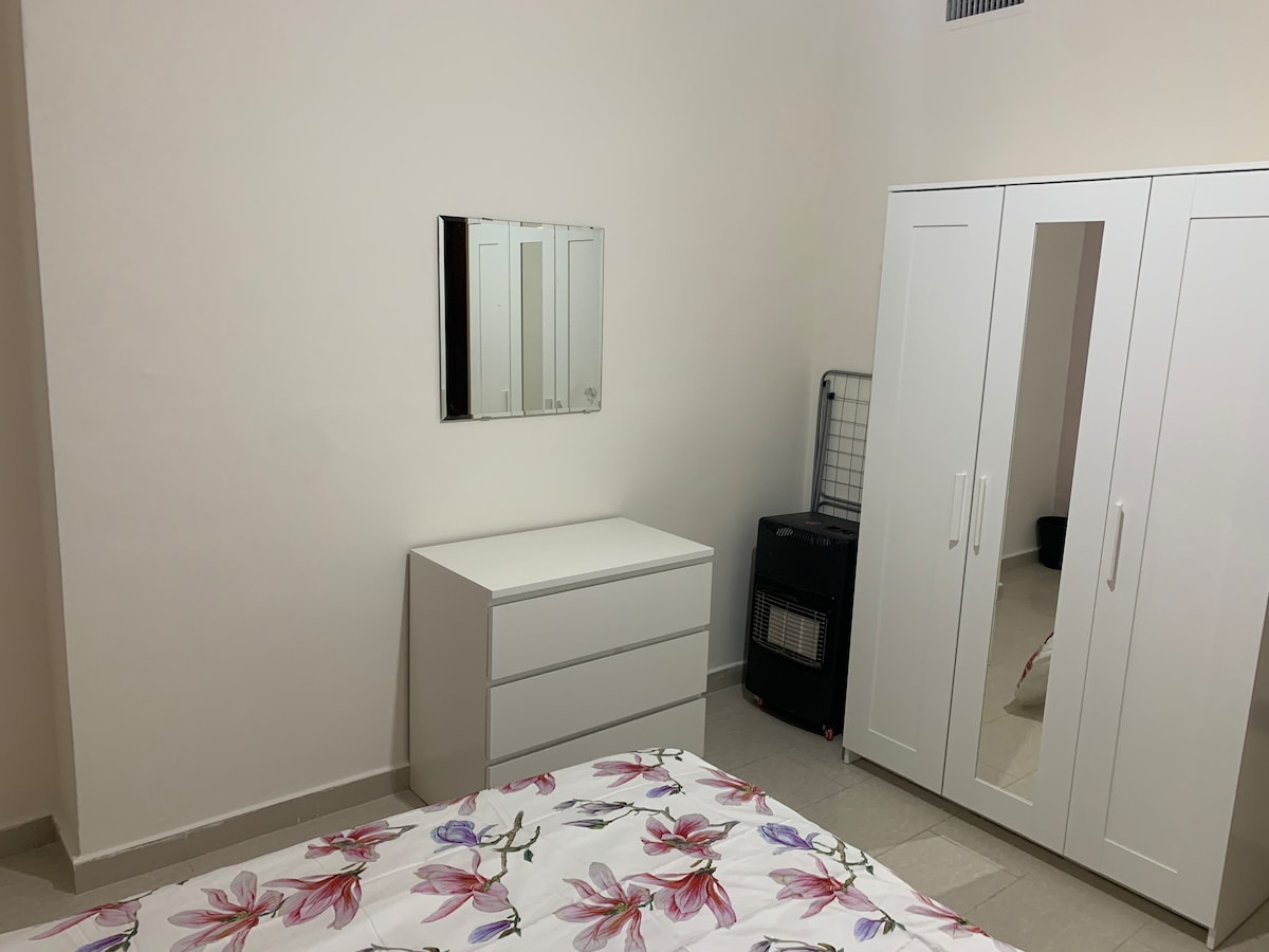 The room features a spacious bed with floral bedding and a modern dresser. A large mirrored wardrobe stands against the wall, while a small storage unit is nearby. Soft lighting enhances the neutral-tone walls and tiled floor.
