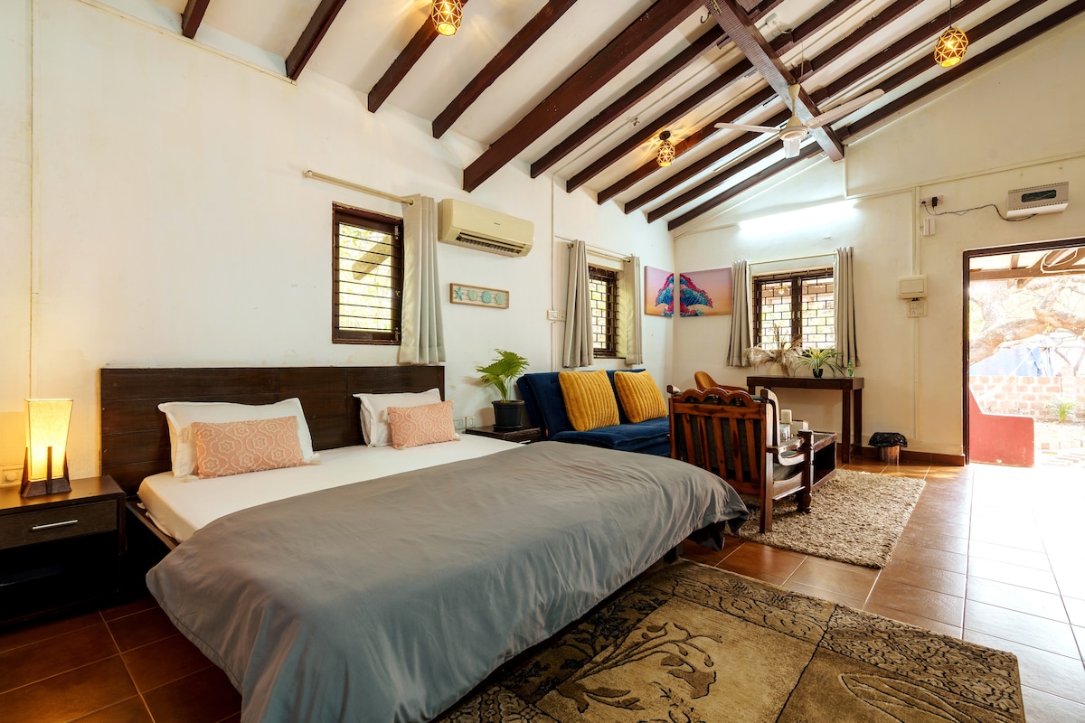 The spacious living area features a large bed with soft bedding alongside a comfortable sofa bed. Accents include wooden beams, earthy tile flooring, and natural light filtering through the windows. A small dining table is also visible, creating a cozy setting ideal for relaxation.