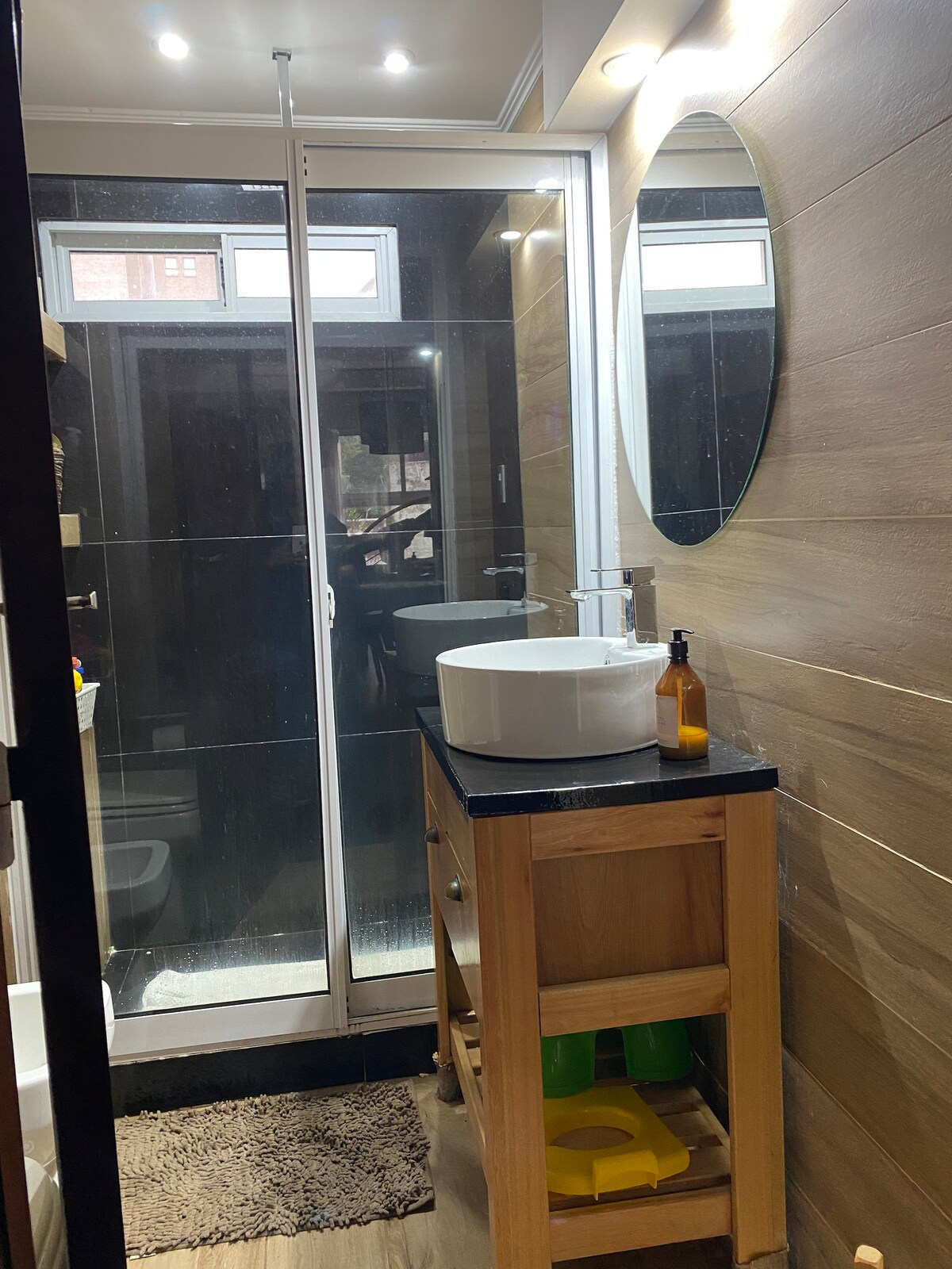 A modern bathroom is illuminated by bright ceiling lights. A circular mirror is mounted above a sleek sink set on a wooden vanity. A glass shower stall with sliding doors is visible in the background, alongside a storage unit featuring colorful items.