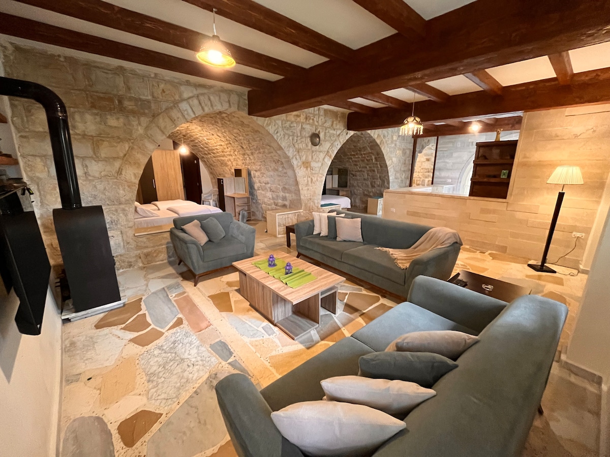 A spacious living area features cozy sofas arranged around a wooden coffee table. Stone walls and wooden beams create a classic atmosphere. Arched openings lead to additional areas, while a textured floor adds character. Soft lighting contributes to a welcoming ambiance.