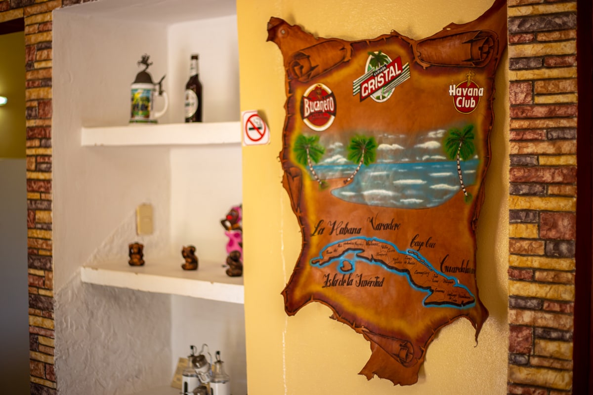 A decorative leather map of Cuba is displayed on a brightly colored wall. The map features illustrations of palm trees and local beverage brands. A set of shelves can be seen nearby, holding various decorative items and a bottle of beer.
