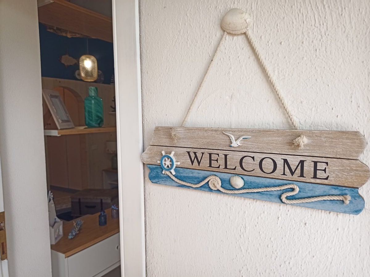 A wooden welcome sign is displayed on a wall, featuring nautical elements such as a seagull and rope details. In the background, a glimpse of a well-organized interior space is visible, with decorative ocean-themed items arranged neatly.
