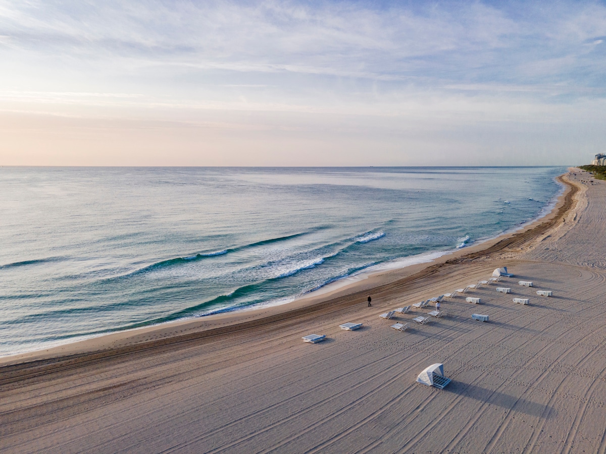 A serene beach stretches along the coastline, with gentle waves lapping at the shore. White umbrellas and lounge chairs are arranged in neat rows on the sand, creating a laid-back atmosphere. The sky displays soft pastel hues as the sun rises over the tranquil water.