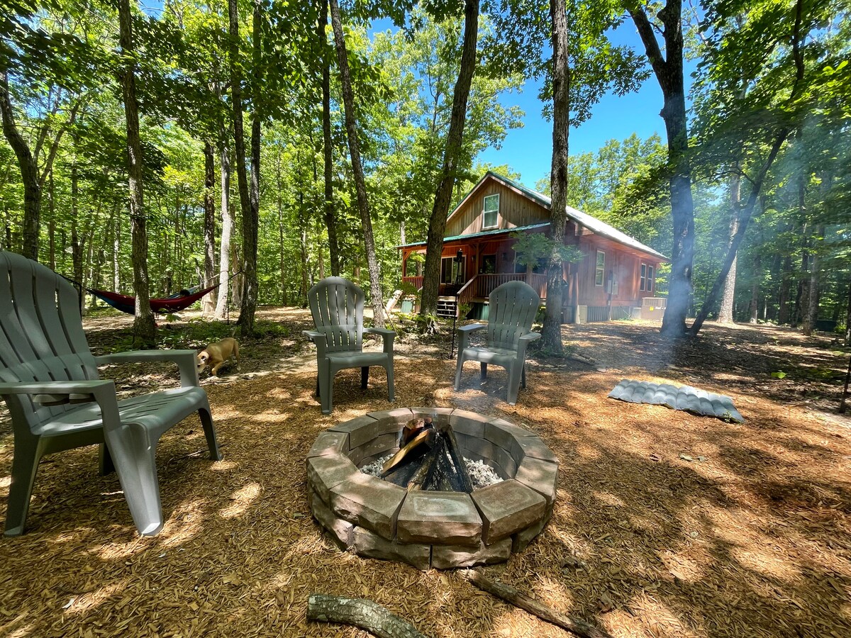 A fire pit surrounded by five gray chairs is set within a wooded area, offering a serene outdoor gathering space. Sunlight filters through the lush tree canopy, illuminating a cabin in the background. A hammock is visible nearby, enhancing the tranquil environment.