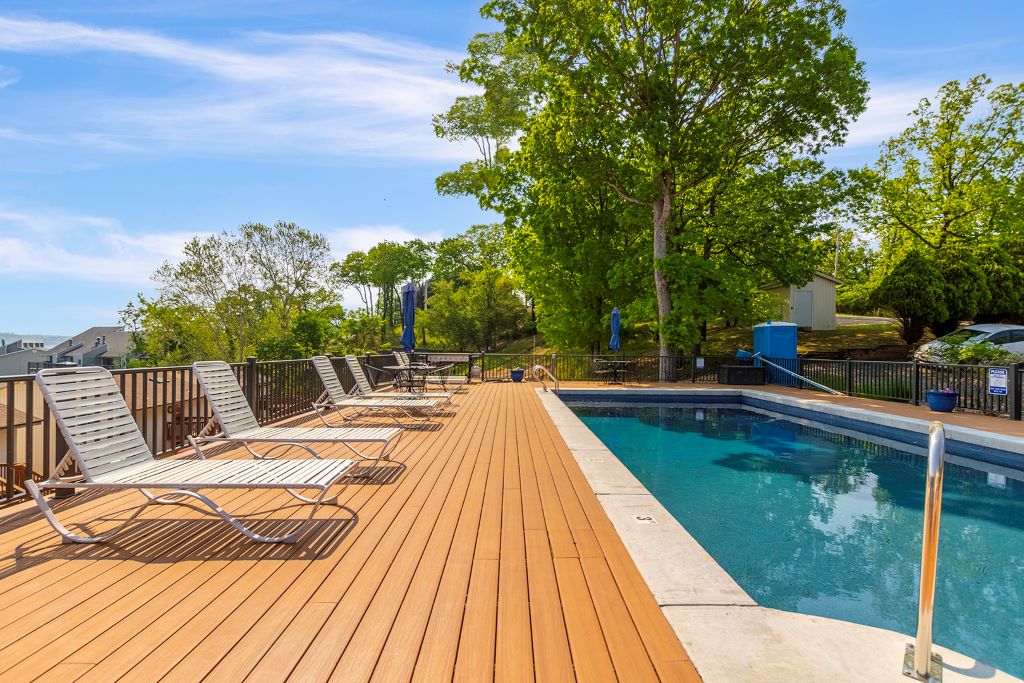 A sunlit pool area is framed by wooden decking, offering several lounge chairs arranged at the edge. Lush greenery provides a natural backdrop, while a gentle breeze ripples across the water's surface. The space invites relaxation and enjoyment of the outdoor surroundings.