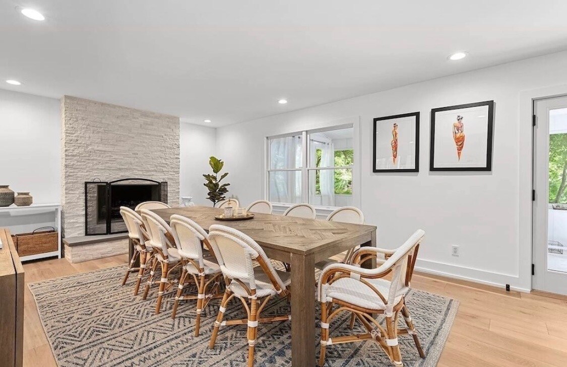 A formal dining area is displayed with a large wooden table surrounded by ten rattan chairs. Light pours in through large windows, highlighting the white decorative stone fireplace and artworks on the walls. A cozy area rug complements the space.