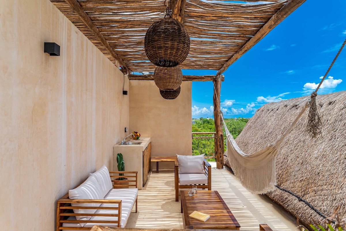 An inviting terrace features natural wood furnishings, including a sofa with white cushions and a wooden coffee table. A hammock is suspended nearby, while the space is shaded by woven overhead decor. A small kitchenette is positioned against the wall, with a scenic view of greenery in the background.