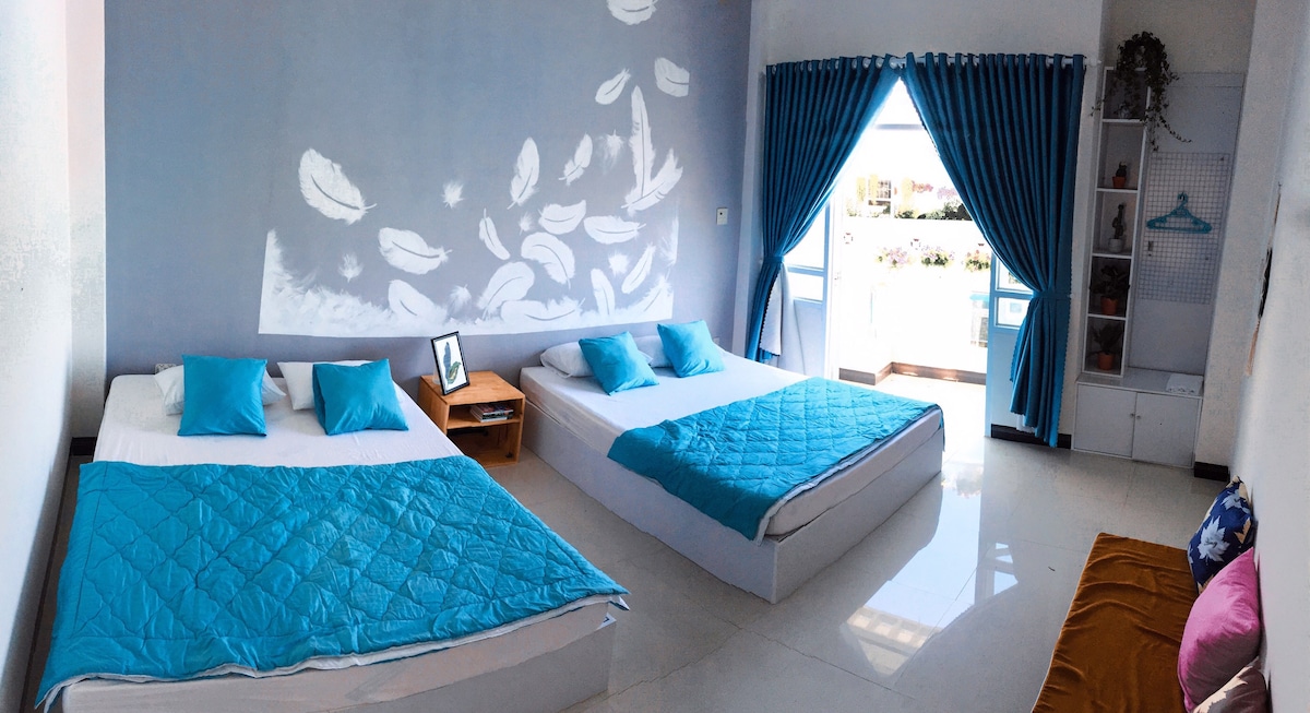 A spacious room features two beds dressed in blue and white bedding. Soft pillows complement each bed. A large window framed by blue curtains allows natural light to fill the room, while a cozy seating area is visible in the corner.