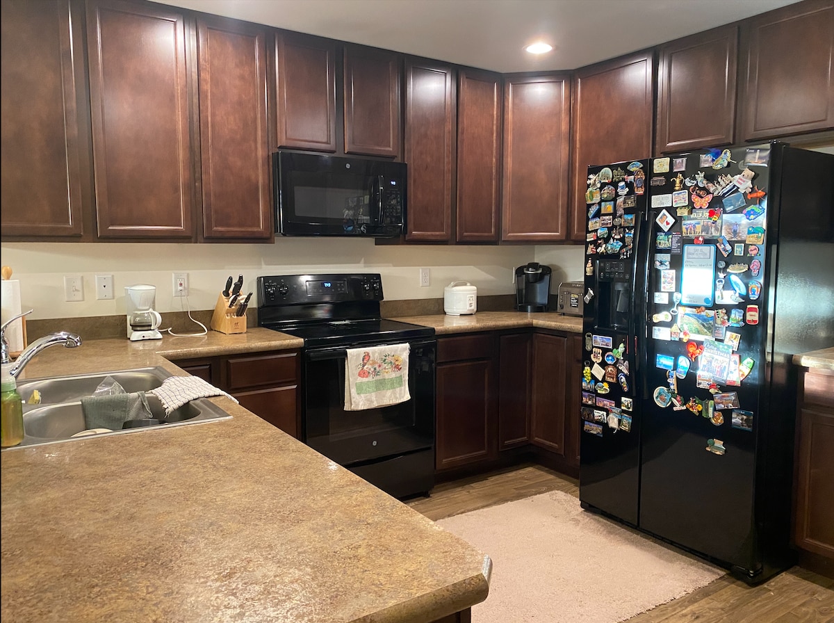 The kitchen features dark wood cabinets and countertops, equipped with modern appliances including a black refrigerator covered in magnets. A stainless-steel sink is positioned beside the counter. Soft lighting enhances the spacious layout, creating a functional cooking area.
