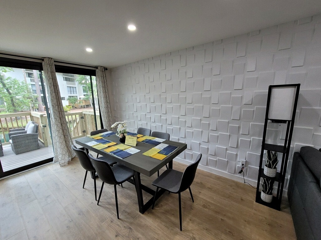 A modern dining area features a large table set with a multi-colored table runner. Six chairs surround the table, and a decorative shelf stands nearby. Large glass doors provide access to an outdoor space, with natural light illuminating the textured wall.