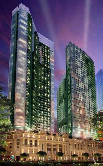Two tall, modern high-rise buildings feature extensive glass facades that reflect the evening sky. The lower level displays a classic architectural design with trees lining the entrance, suggesting a blend of contemporary and traditional styles.