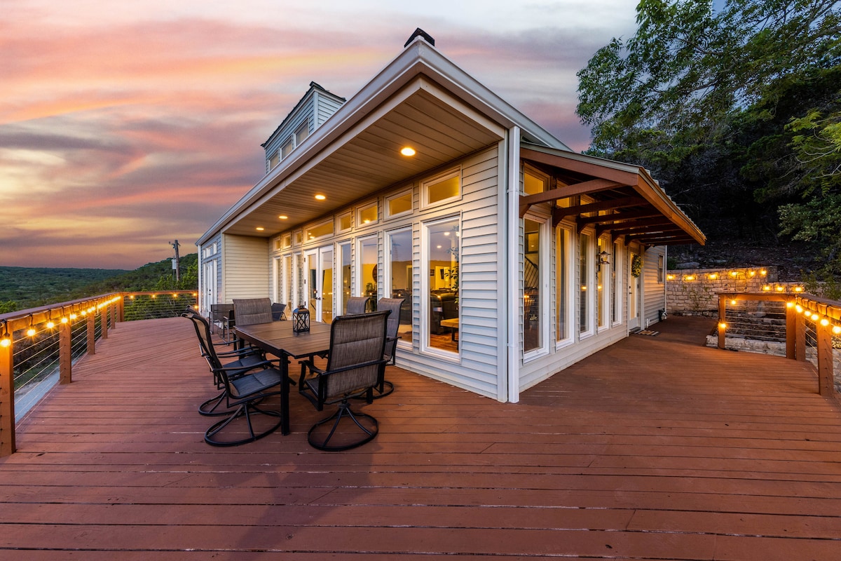 A large wraparound deck features multiple seating arrangements and a dining table, illuminated by string lights. Expansive windows provide stunning views of the surrounding landscape, while the sunset casts a warm glow over the home, enhancing the outdoor space.
