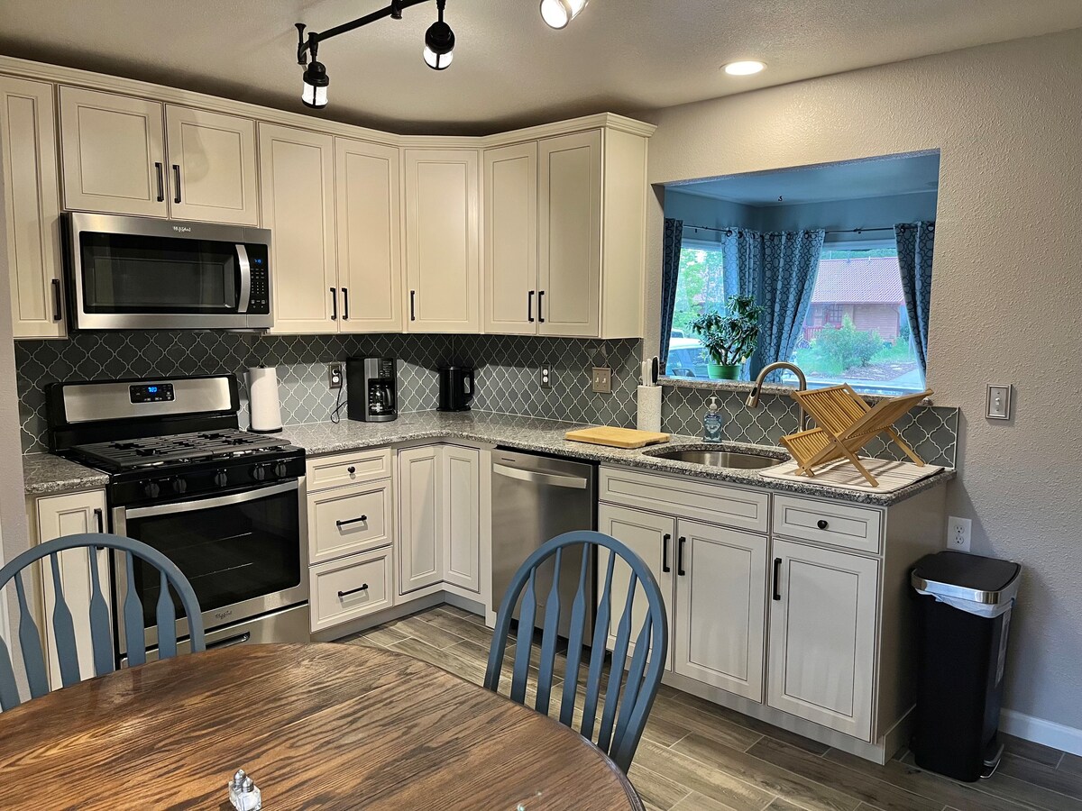 The kitchen features modern white cabinetry and a stylish backsplash, complemented by stainless steel appliances. A large window allows natural light to fill the space, offering a view of the outdoor greenery. A round wooden table is positioned centrally, surrounded by dark chairs.