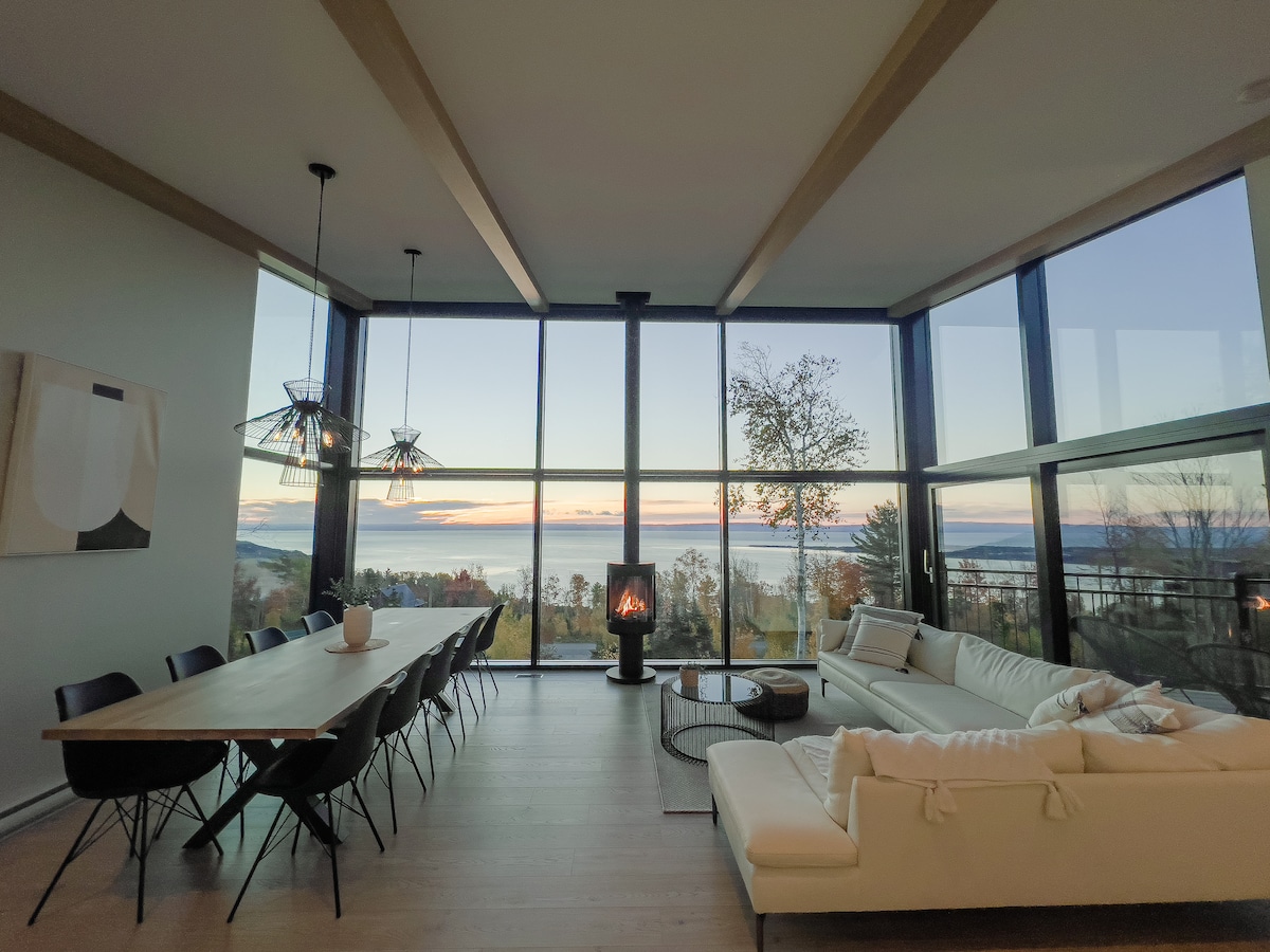 A spacious living area is visible, featuring large floor-to-ceiling windows that offer a panoramic view of the surrounding landscape and water. A modern white sectional sofa and a round coffee table are positioned to invite relaxation. A dining table with black chairs is present, illuminated by stylish light fixtures.