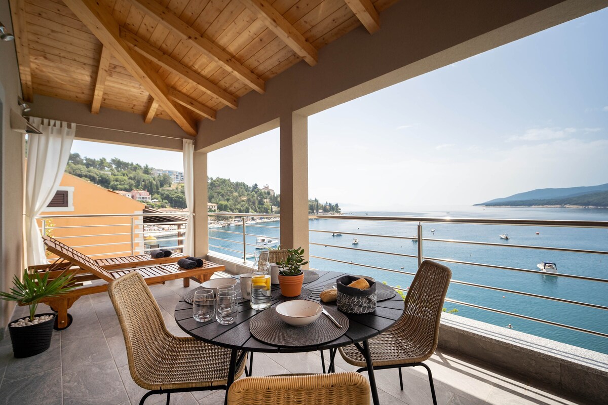 A spacious terrace with a wooden ceiling offers direct sea views. A round table is set for dining, accompanied by four woven chairs. Two sun loungers are positioned nearby. The surrounding area features potted plants, enhancing the serene coastal setting.