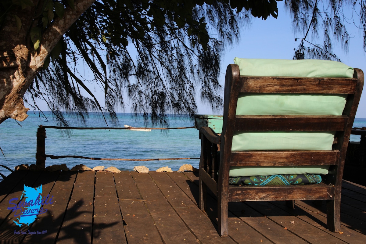 A wooden chair with green cushions is positioned on a wooden deck overlooking the ocean. Sunlight filters through the surrounding foliage, creating a serene environment. The calm water reflects a blue sky, enhancing the natural setting.