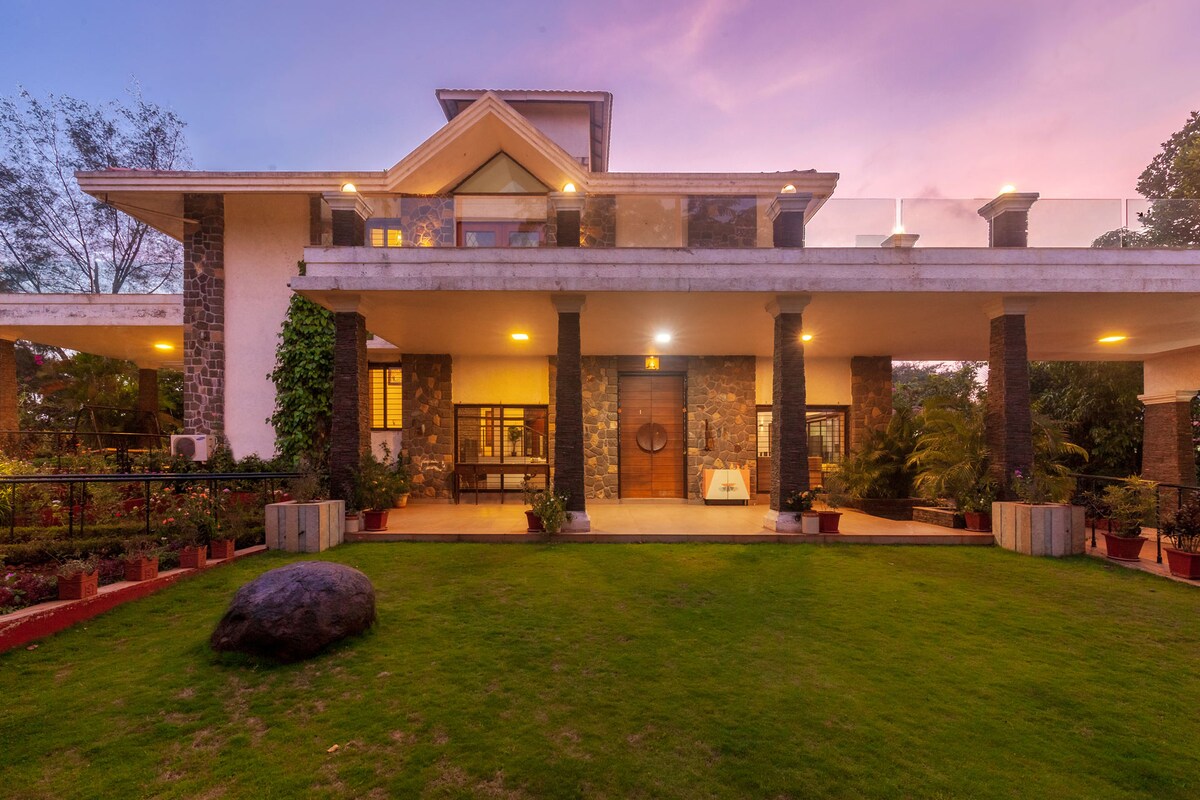 The exterior of the bungalow showcases a two-story structure surrounded by lush green lawns. The stone facade is complemented by large, illuminated windows and a spacious porch, offering an inviting entrance. Soothing evening light casts a warm glow across the property.