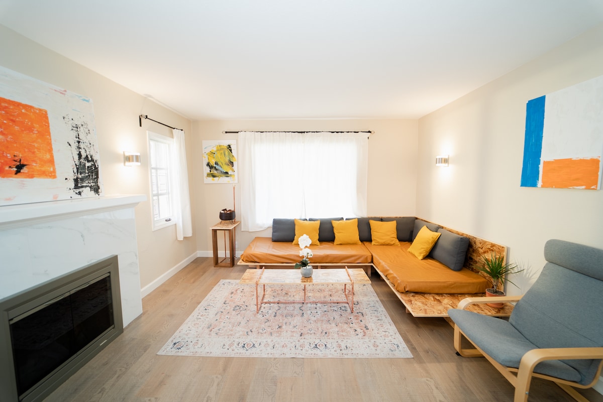 A bright living room features a modern sectional sofa adorned with orange and gray cushions, complemented by a light wooden coffee table. Two art pieces decorate the walls, and a window admits natural light, enhancing the room’s inviting atmosphere. A fireplace is subtly placed to one side.