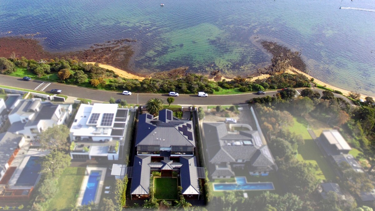 Aerial view highlights the stunning waterfront location of the accommodation, showcasing the beautiful shoreline with visible turquoise waters. Nearby properties are seen, including landscaped gardens and a road offering direct access to the beach.