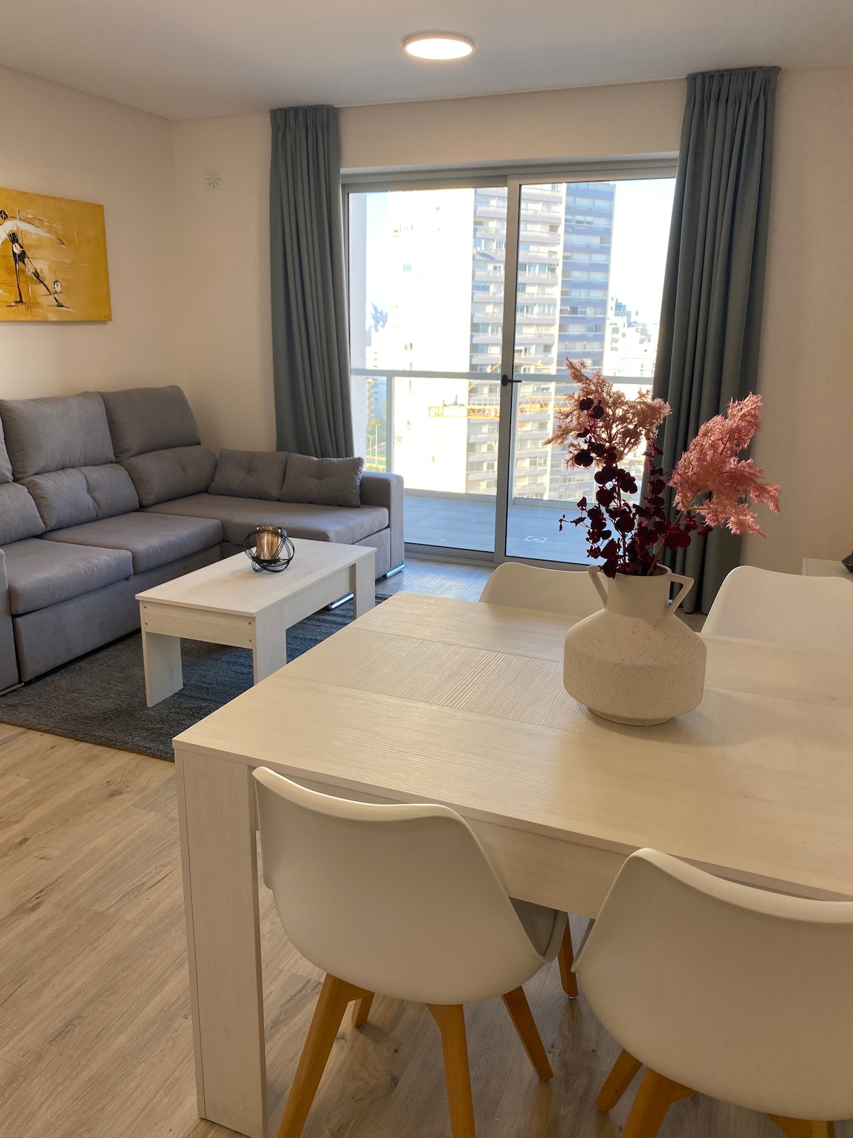 A contemporary living space showcases a gray sofa accompanying a white coffee table, positioned on a light area rug. A dining table with four chairs is visible, and a bouquet in a vase adds a touch of color. Natural light floods the area through large windows.