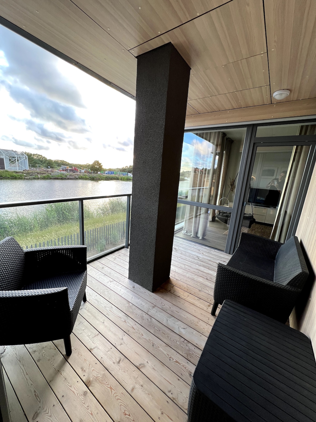 A private balcony features two black wicker chairs and two small matching tables, offering a relaxing space overlooking calm waters. The wooden deck adds a natural element, while large glass doors lead to the interior, allowing for easy access and stunning views.
