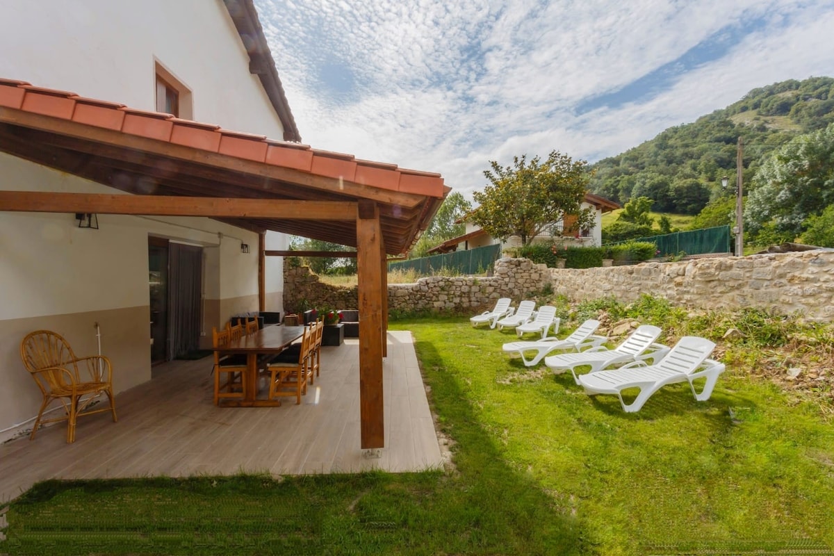 A gently shaded outdoor area features a long wooden table accompanied by several light-colored chairs. Sun loungers are arranged on the grassy lawn, with a stone wall and greenery visible in the background. A view of a hillside is present, enhancing the serene environment.