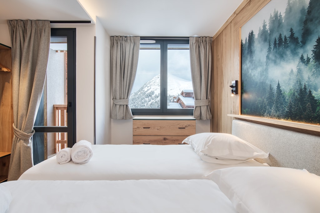 A well-appointed guest room features two spacious beds with neutral bedding, complemented by a scenic wall mural of trees and mountains. Large windows allow natural light to fill the space, while soft curtains frame the views of the surrounding landscape.