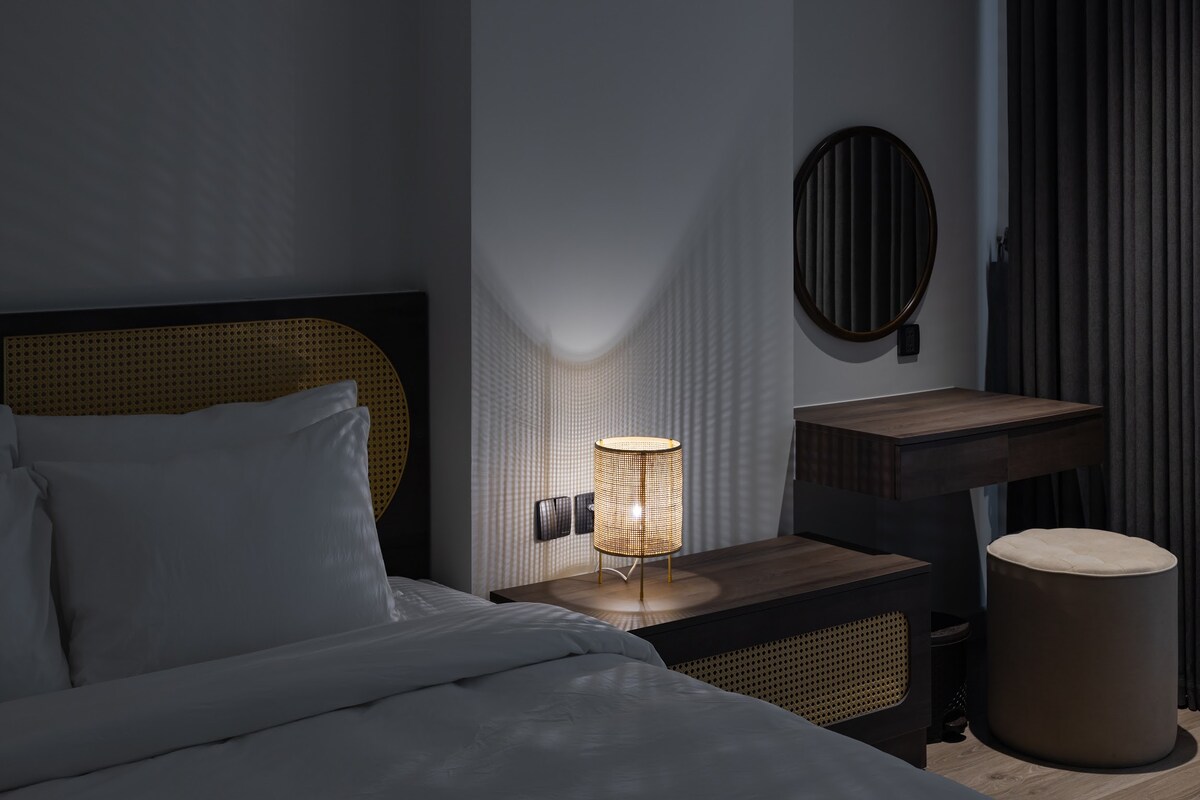 A restful bedroom is displayed with a spacious bed featuring crisp white linens. A soft glow emanates from a woven bedside lamp, casting gentle shadows. A round mirror and a simple wooden table are positioned nearby, complemented by a small, upholstered stool.