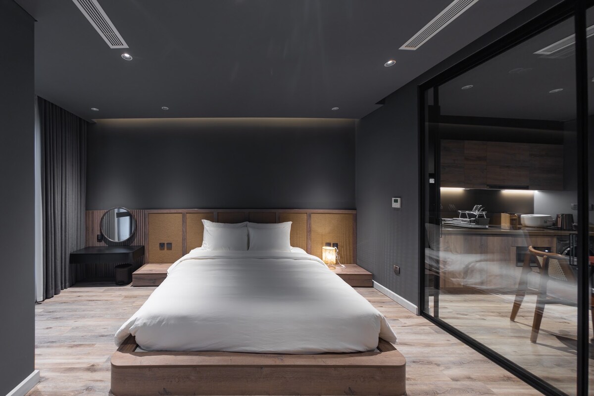 A spacious bedroom features a large, minimalistic bed with a white duvet. Natural light enters through a glass wall that separates the sleeping area from the kitchen. Soft tones are complemented by wooden flooring and accents, creating a calm and stylish environment.