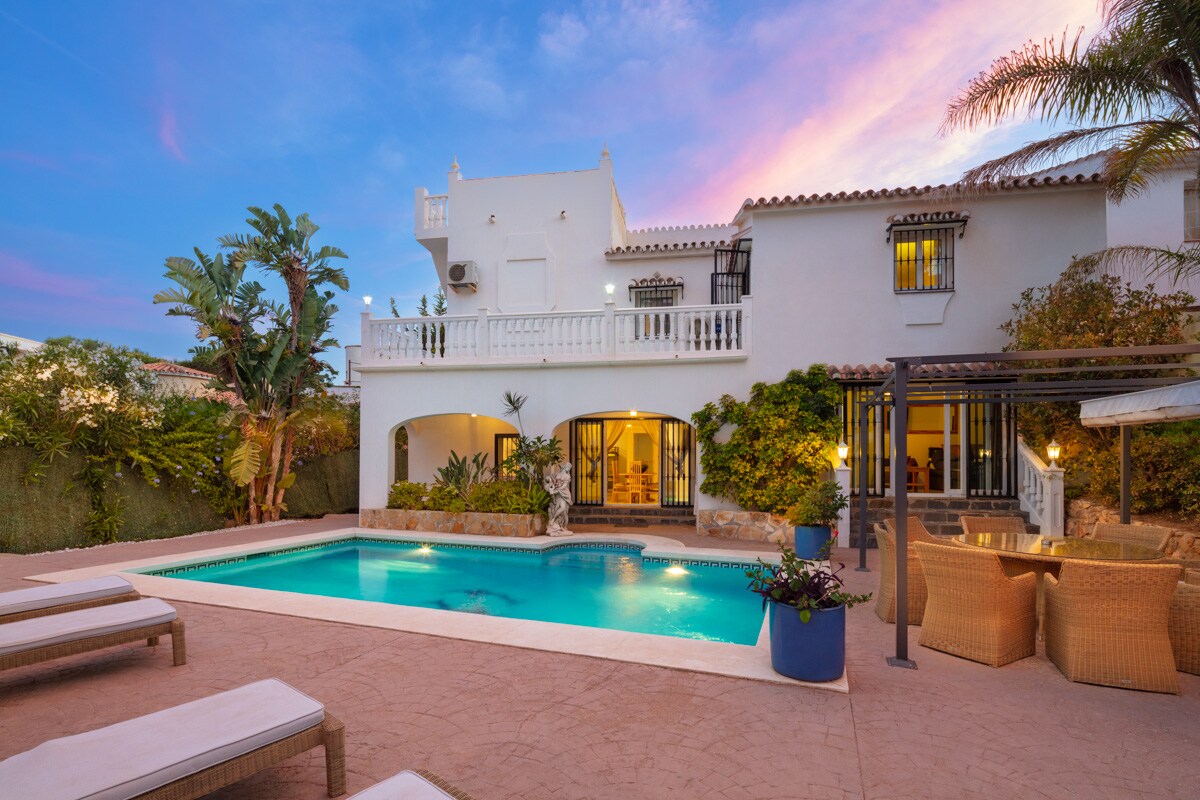 The two-story villa features a private swimming pool surrounded by a sun terrace with comfortable lounge chairs. Lush greenery and palm trees frame the outdoor area. Large glass doors invite natural light into the living spaces visible from the patio, enhancing the open and inviting ambiance.
