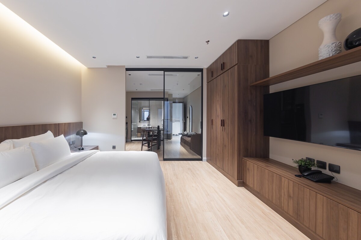 A modern bedroom features a spacious bed with crisp white linens, complemented by warm wooden accents. A large glass door leads to an adjacent space, while a flat-screen TV and minimalistic shelving are present. Soft lighting enhances the serene atmosphere.