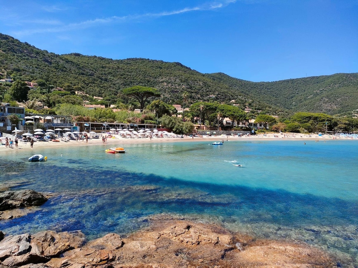A sandy beach stretches along a clear turquoise sea, with gentle waves lapping at the shore. Umbrellas and sun loungers are set up for visitors, while rocky outcrops provide texture to the coastline. Lush green hills rise in the background, creating a serene natural setting.