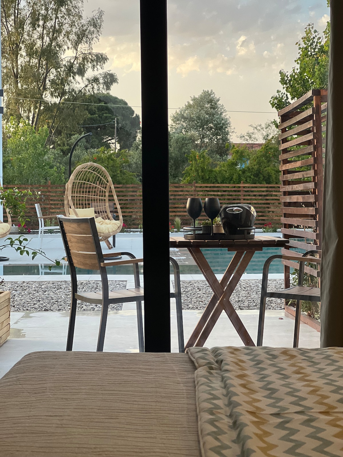 A view from the interior showcases a cozy seating area with a wooden table set for two, featuring glasses and a coffee maker. Outside, a spacious swimming pool is visible, surrounded by greenery and lounge chairs, framed by a large glass door.