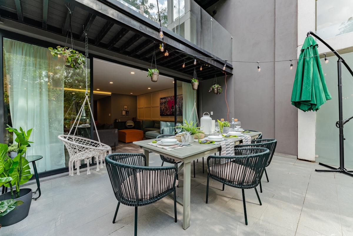 An outdoor terrace features a large dining table set for six, surrounded by four accent chairs. Green accents from hanging plants and a large umbrella complement the space. Soft lighting is provided by string lights, creating a welcoming atmosphere adjacent to a glass wall.