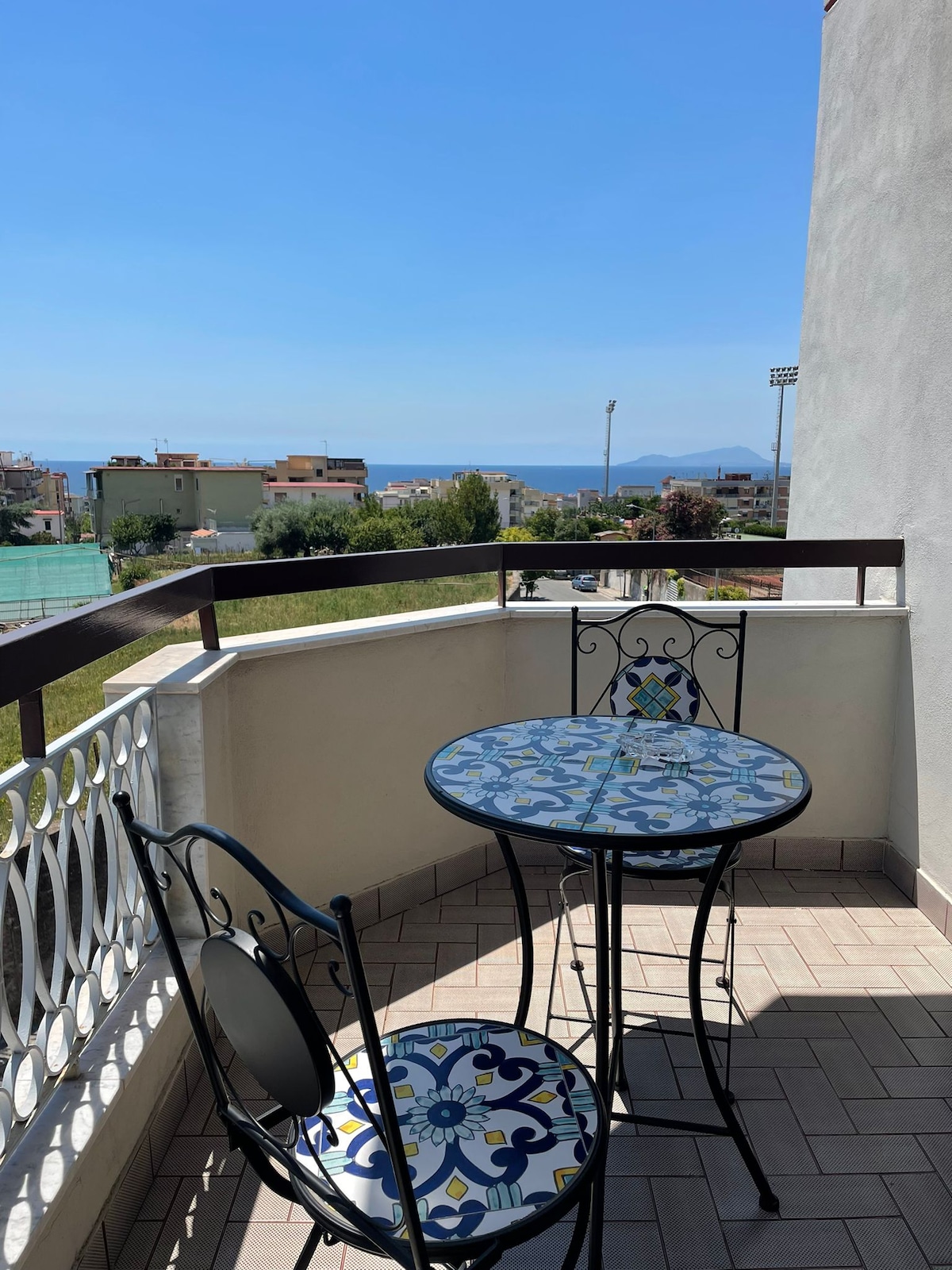 A small balcony features a round table adorned with colorful tile patterns and two black metal chairs. The expansive view reveals distant waters and greenery, with a clear blue sky above. The area is surrounded by buildings and sports facilities, creating a peaceful outdoor space.