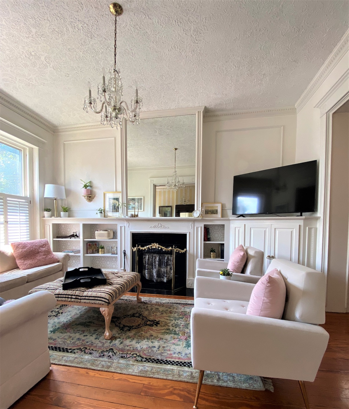 The living area showcases a light-colored seating arrangement with plush white chairs, accented by pink cushions. A decorative area rug is placed on hardwood floors. A mirrored wall reflects natural light, enhancing the space, while a television is mounted nearby above a classic fireplace.