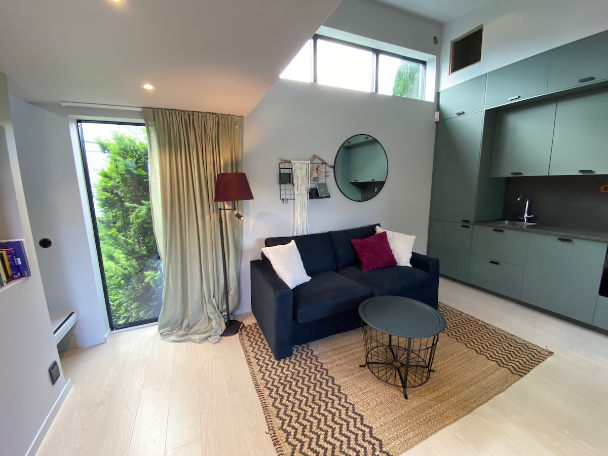 A cozy living area features a dark blue sofa with decorative pillows, a round black coffee table, and a textured area rug. Natural light enters through large windows, creating an open feel. A modern lamp and a circular mirror add to the minimalist decor.